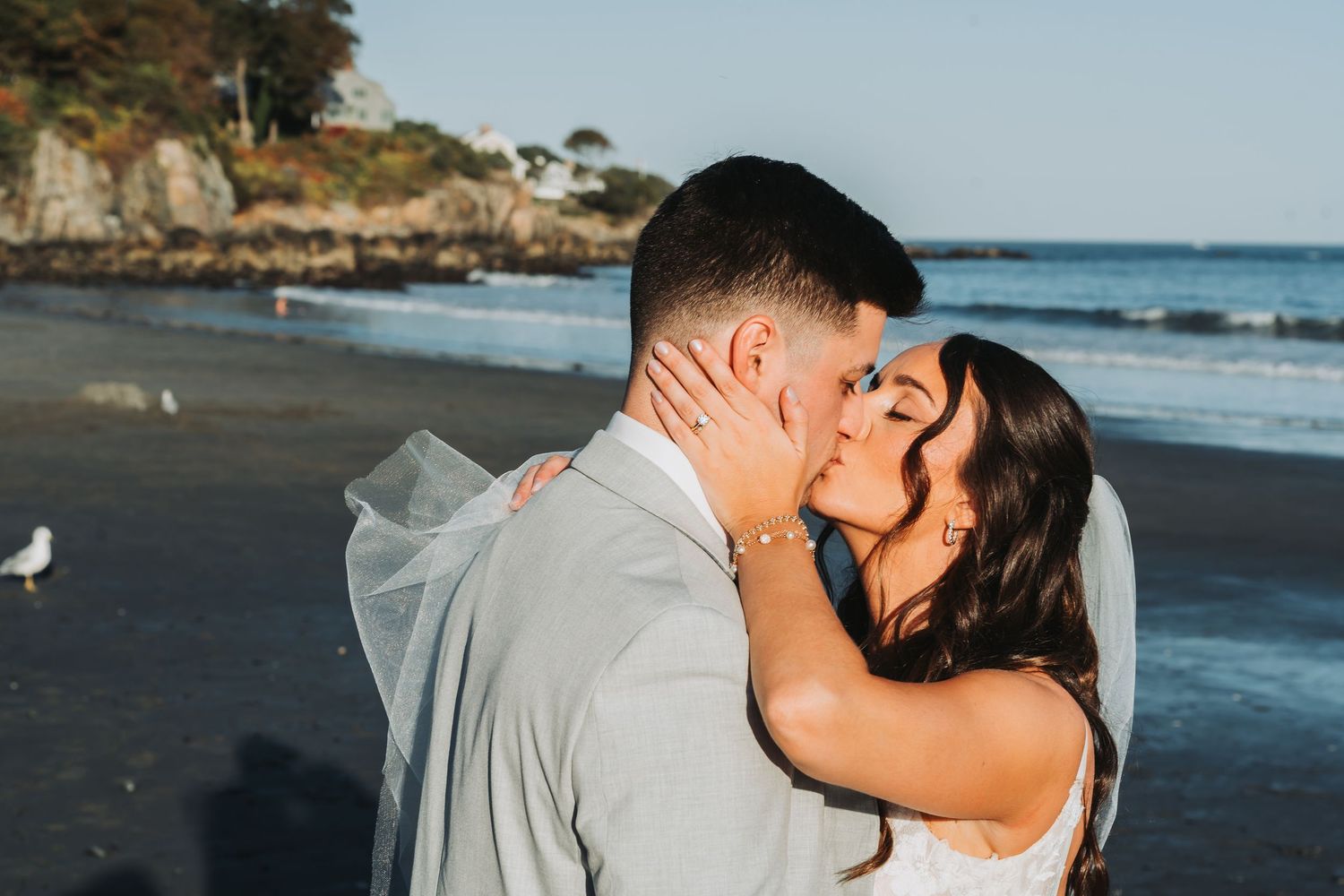 Bride and groom share a romantic beachside kiss at sunset with ocean waves and rocky cliffs behind them.