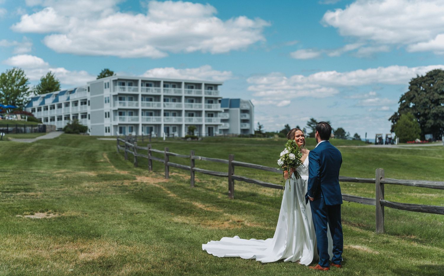 Bride and groom stand by a wooden fence on lush green grounds with a elegant white hotel building backdrop.