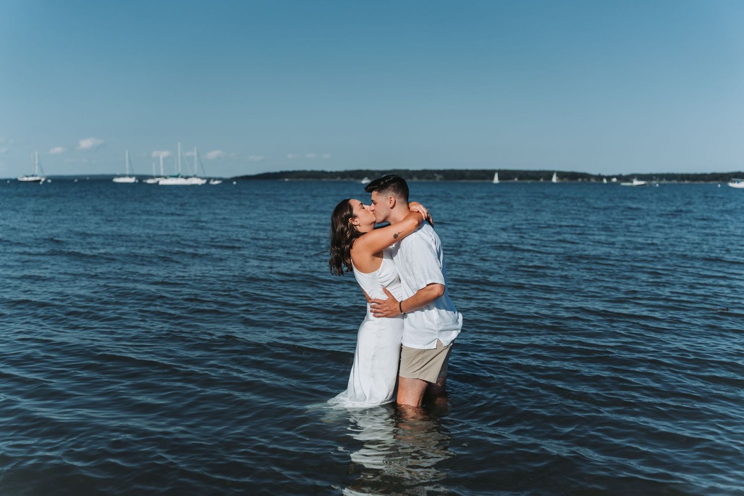 Couple kissing while standing in ocean water during romantic beach engagement photo session on a sunny day.