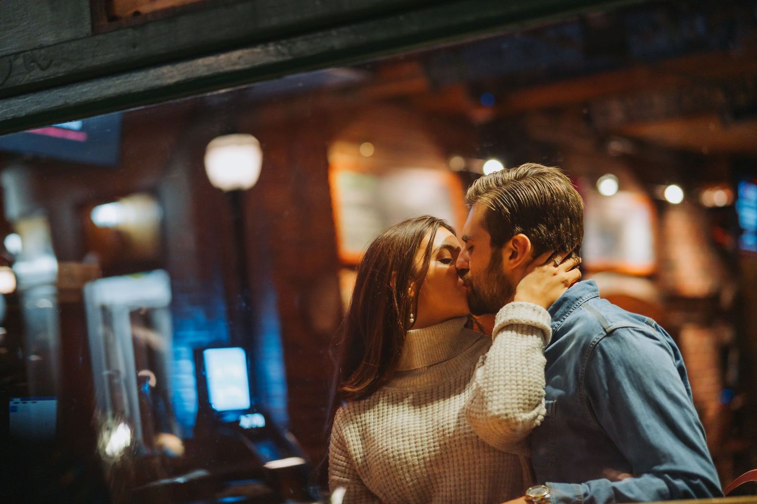 A couple sharing a romantic kiss in a warmly lit bar, the woman in a cozy sweater and man in a blue shirt.