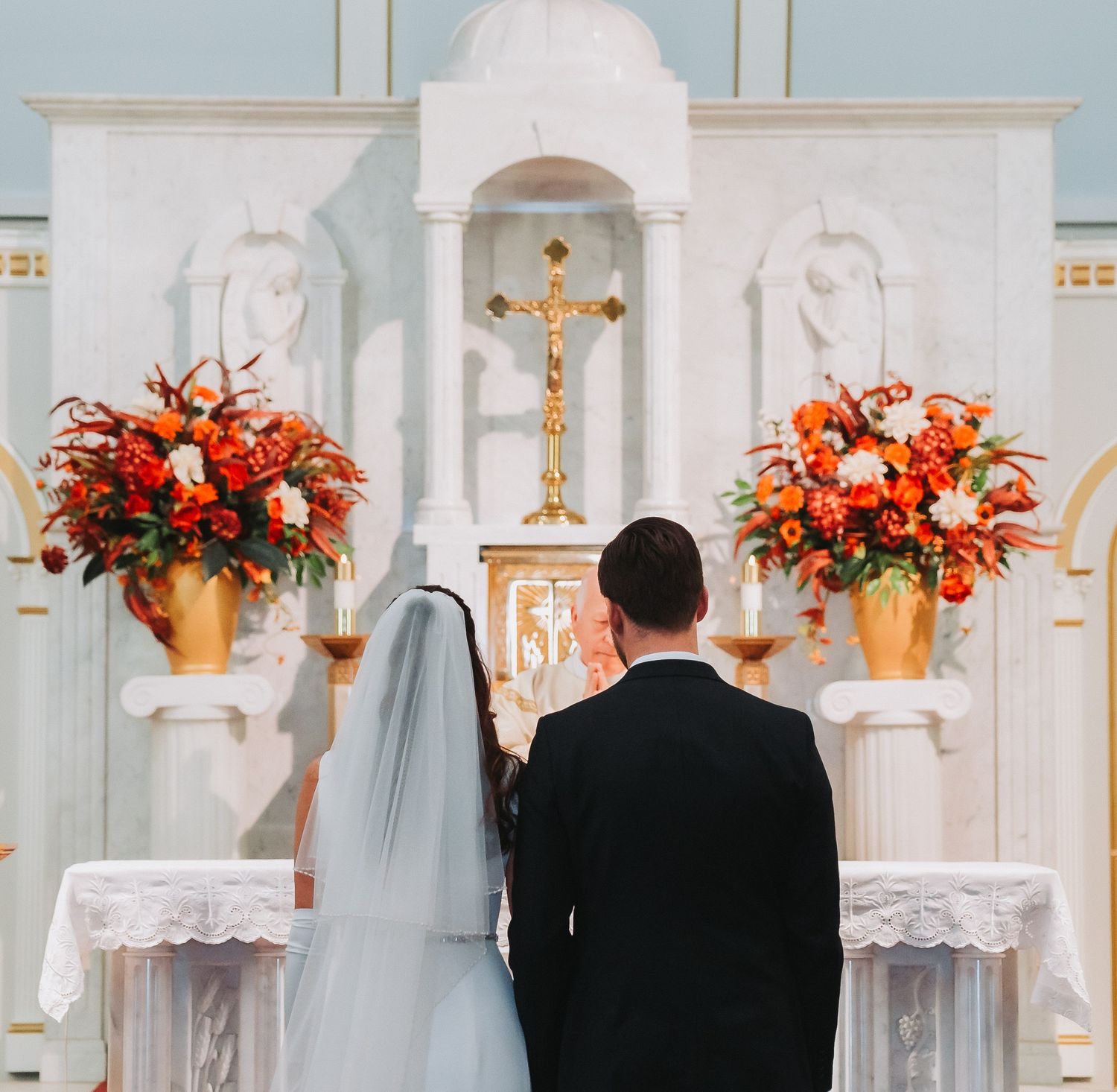 Bride and groom standing at ornate church altar adorned with vibrant orange floral arrangements.
