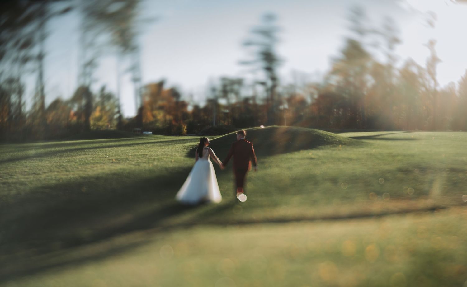A bride and groom walk hand in hand across a sunlit green field surrounded by autumn trees.
