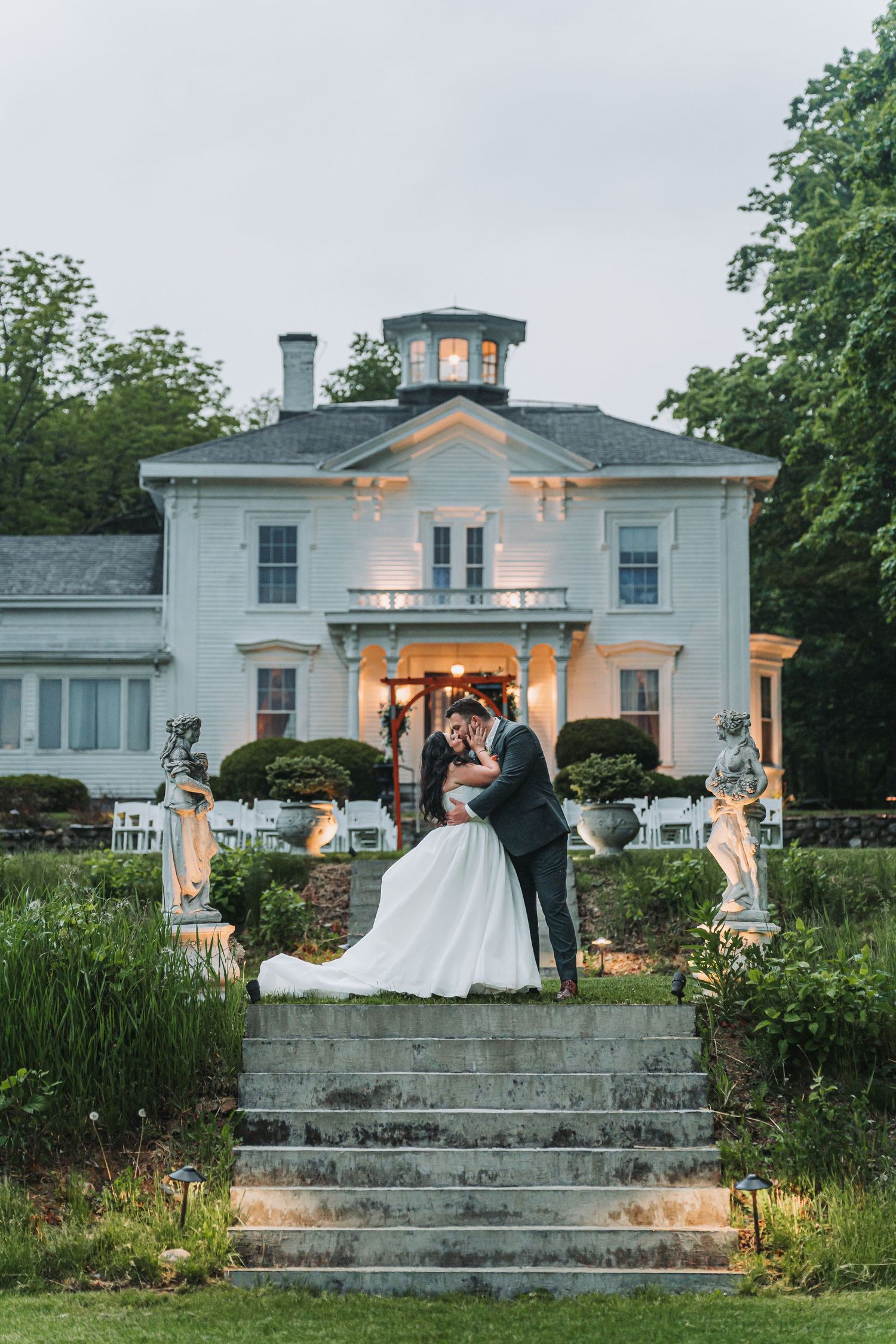 Bride and groom share a romantic embrace on stone steps before an elegant white mansion at dusk.
