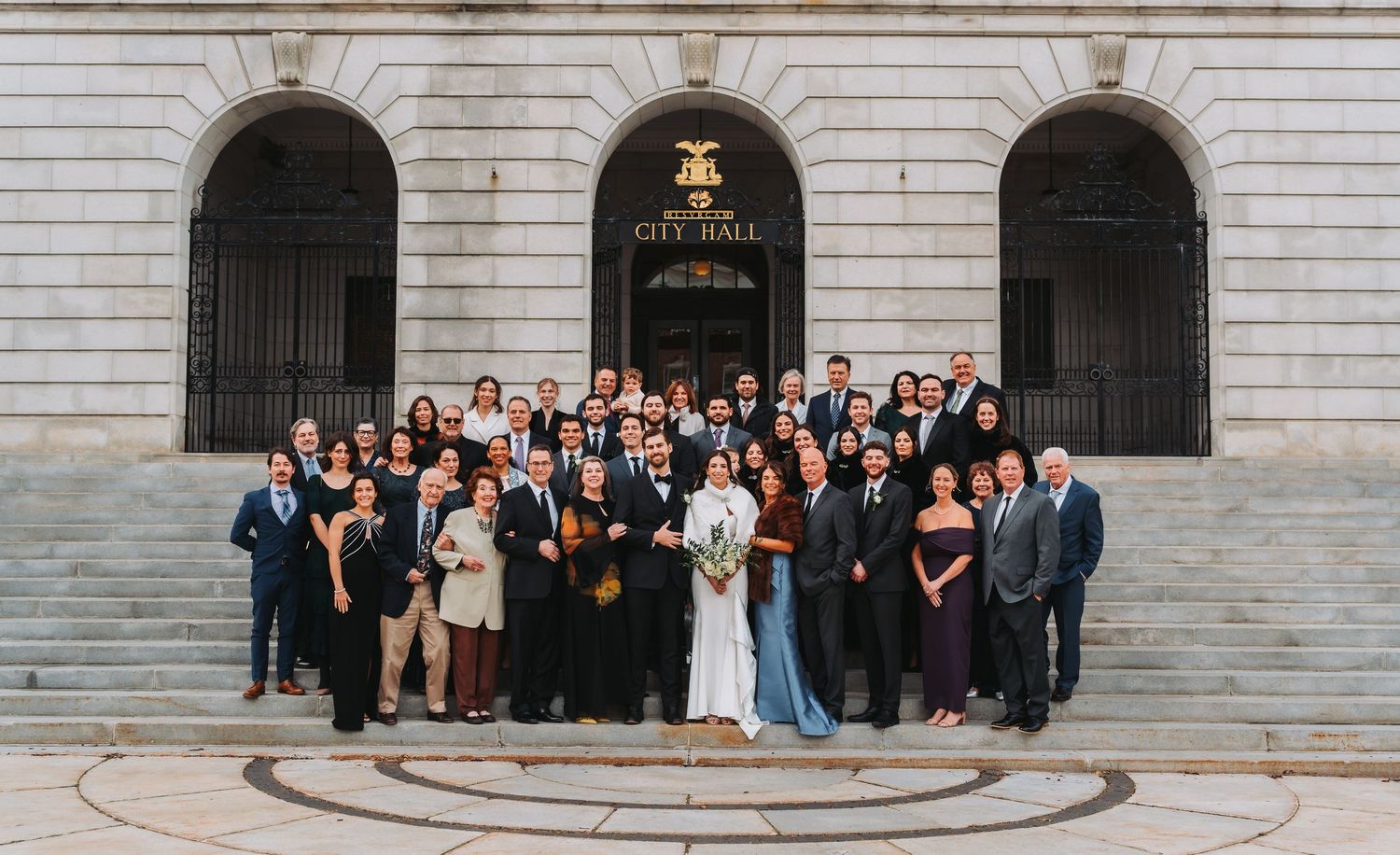 Large wedding party group photo on steps of historic City Hall building after ceremony.