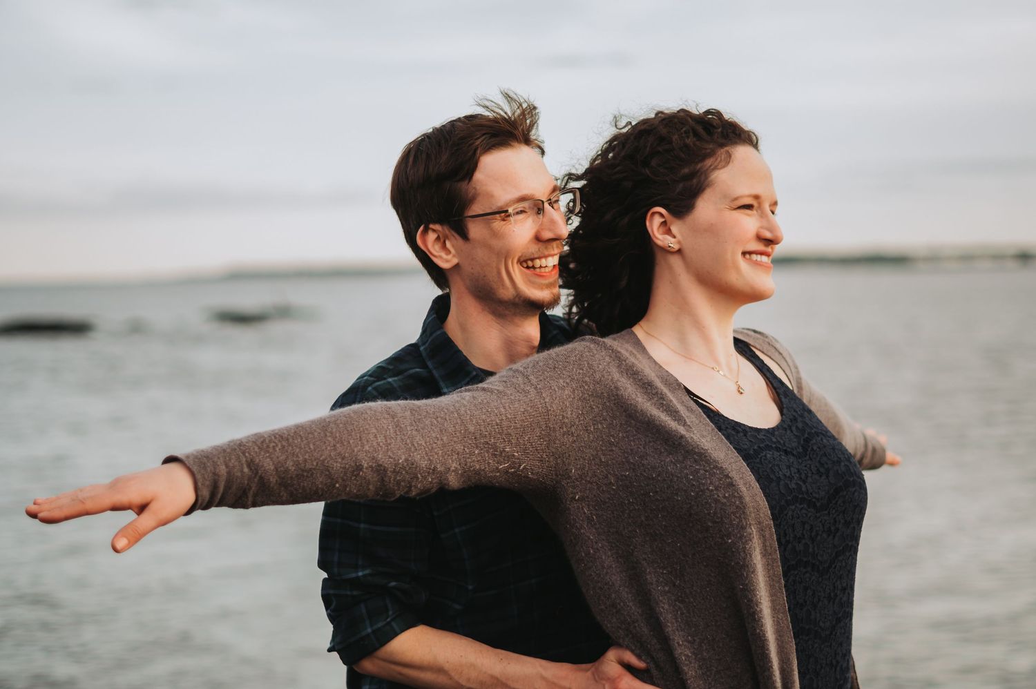 Smiling couple posing with arms outstretched by the ocean at sunset.