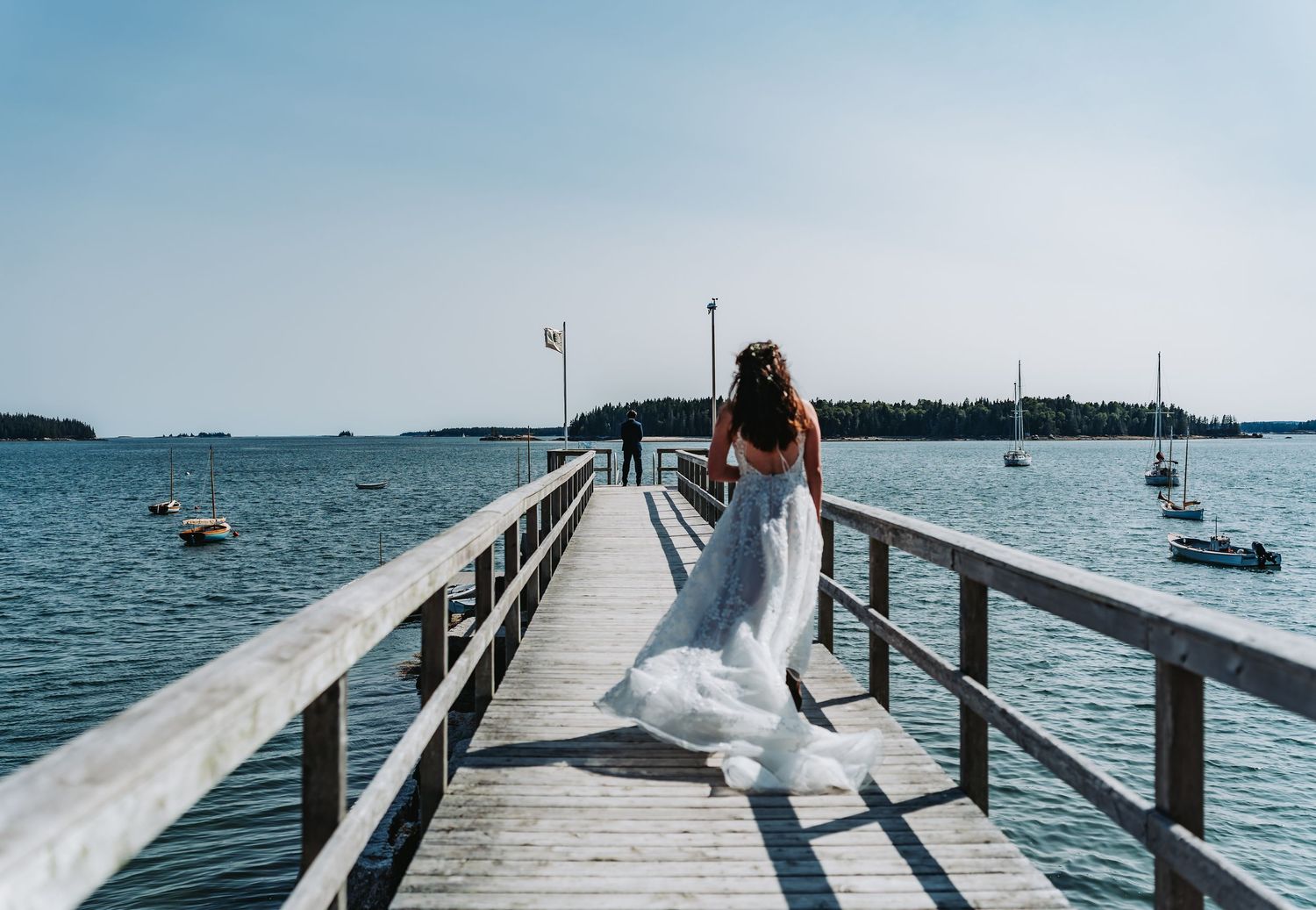 Bride in white gown walks along a wooden pier overlooking a serene lake with sailboats on her wedding day.