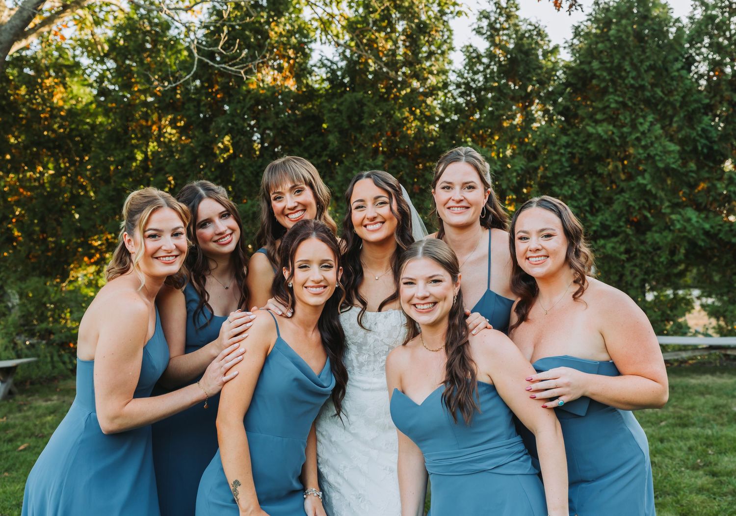 Bride surrounded by seven bridesmaids in blue dresses smiling outdoors in a garden setting.