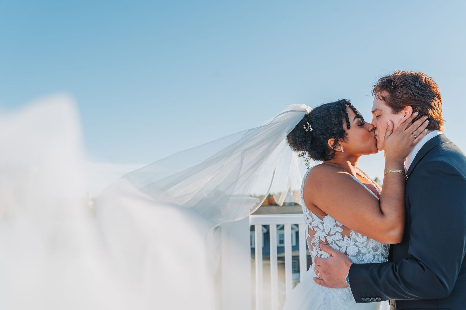 Bride and groom share a romantic kiss as her veil blows in the wind against a blue sky.