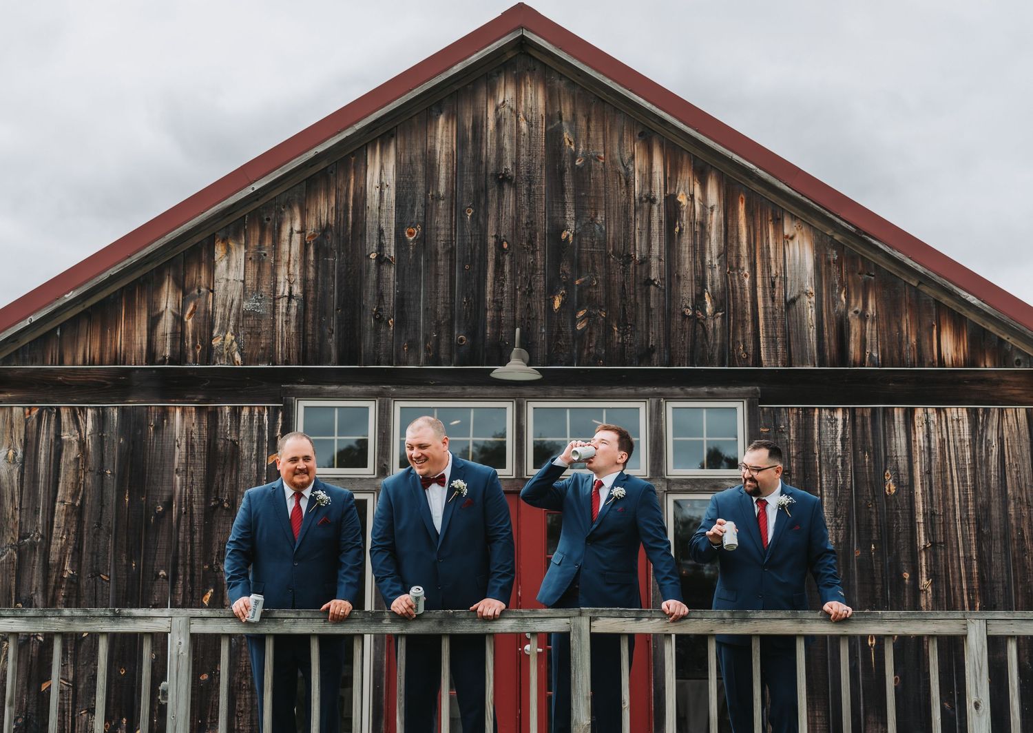 Four groomsmen in navy suits laughing on rustic barn balcony during outdoor wedding celebration.