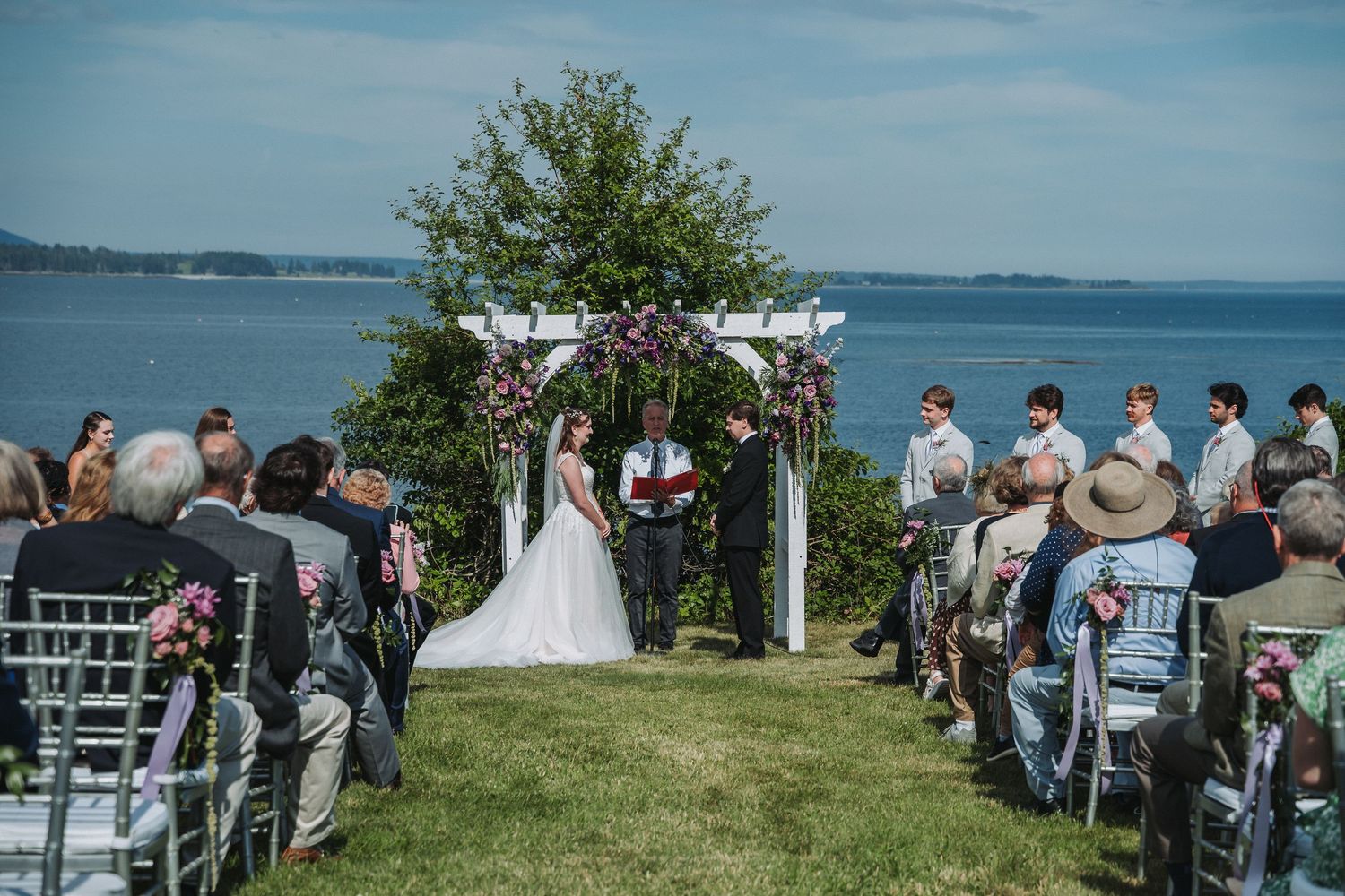 Outdoor waterfront wedding ceremony with couple at floral arch, guests seated on lawn overlooking scenic bay.