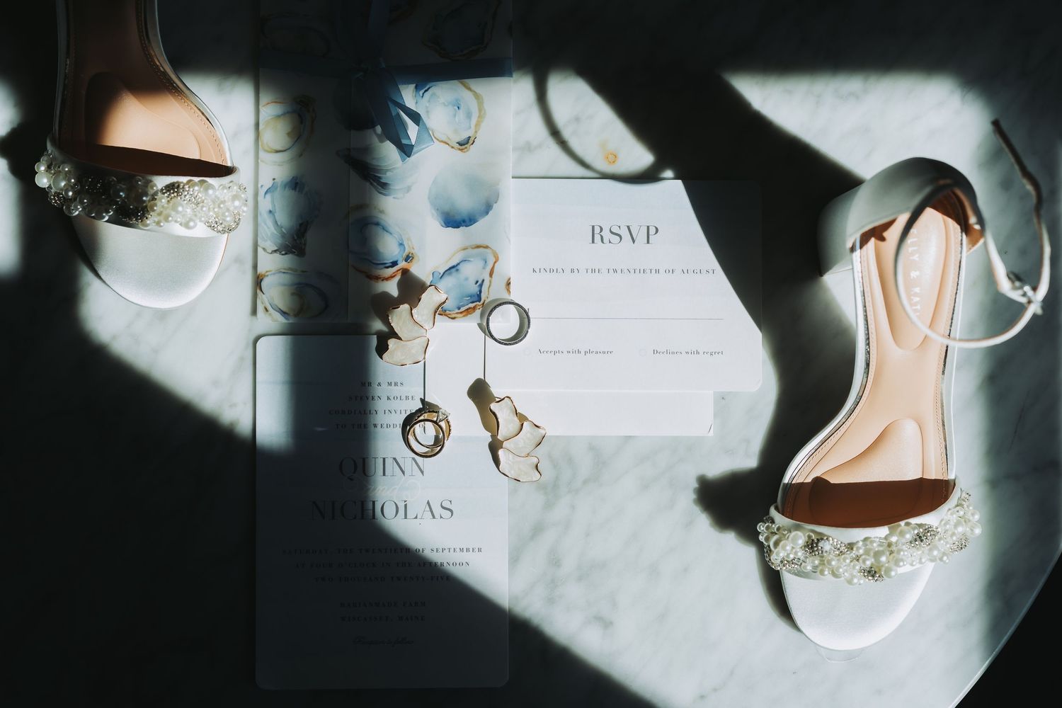 White bridal heels with pearl details displayed alongside wedding invitations and gold earrings in dramatic sunlight.