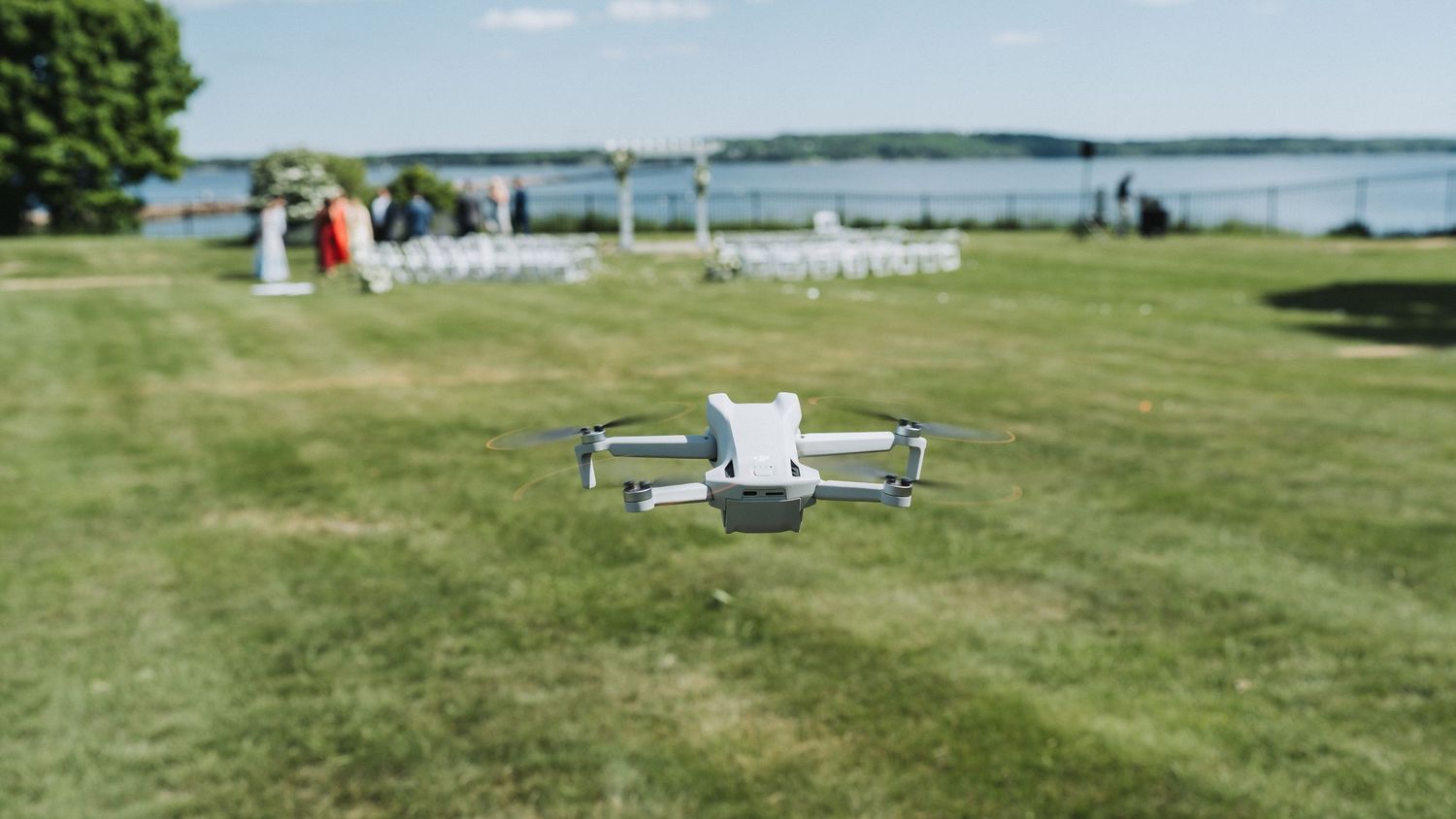 A DJI drone hovering over a green lawn near a waterfront outdoor wedding ceremony setup on a sunny day.