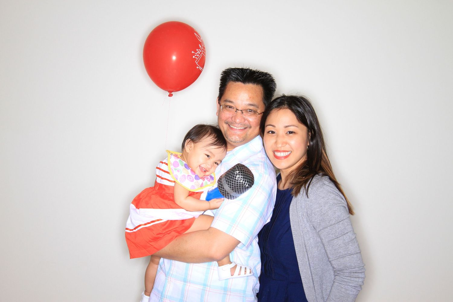 A happy Asian family of three poses with a red balloon, parents smiling while holding their young child.