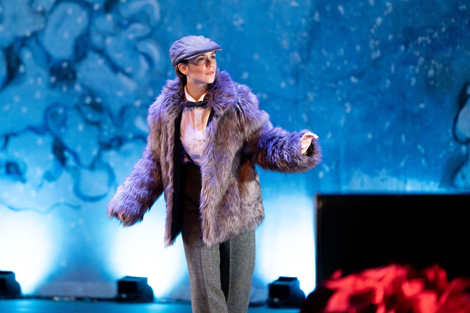 Performer on stage wearing a purple fur coat and newsboy cap under blue dramatic lighting at Las Positas College in Livermore.
