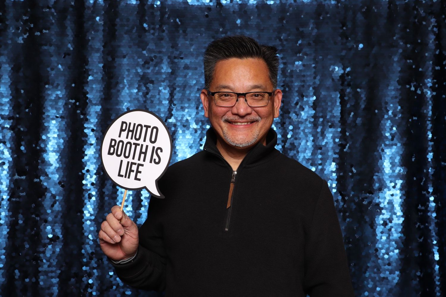Man smiling in photo booth holding a speech bubble sign reading Photo Booth Is Life against a blue sequin backdrop.