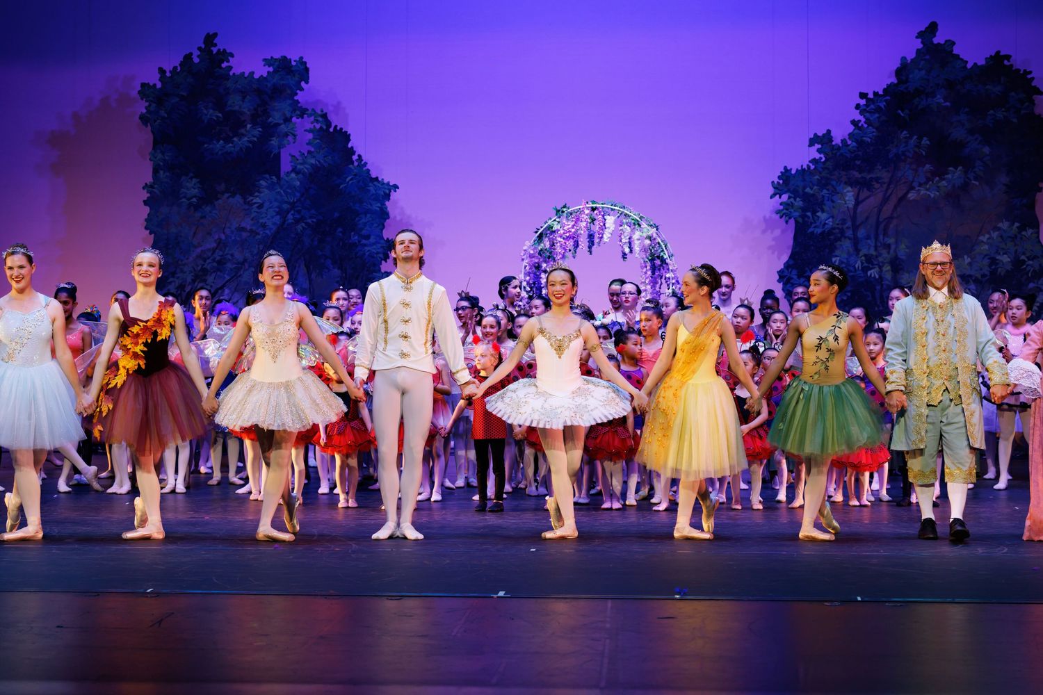 Ballet dancers in colorful costumes take a bow on stage during a performance finale under purple stage lighting.