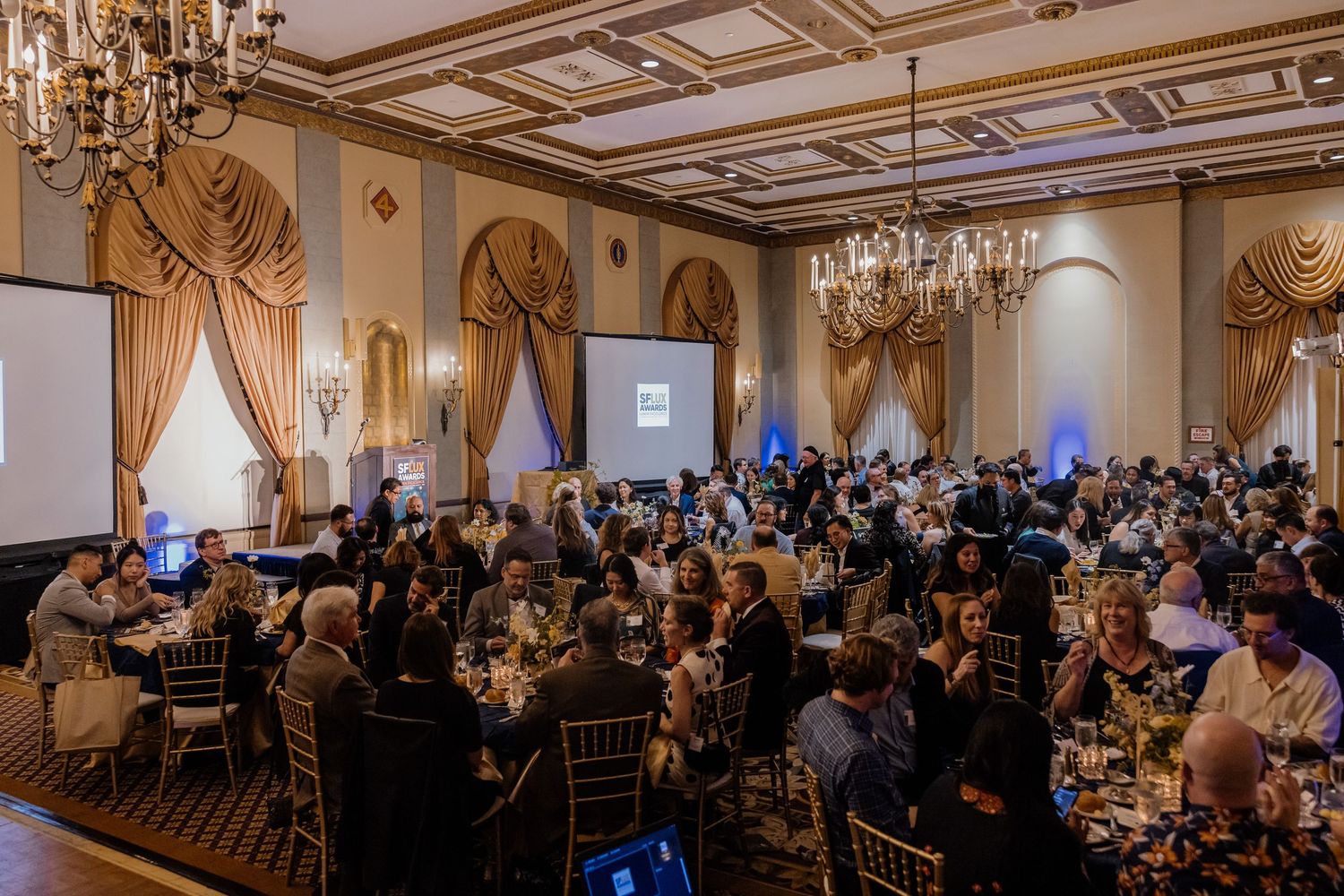 Elegant ballroom gala event with guests seated at round tables beneath chandeliers with a projection screen.