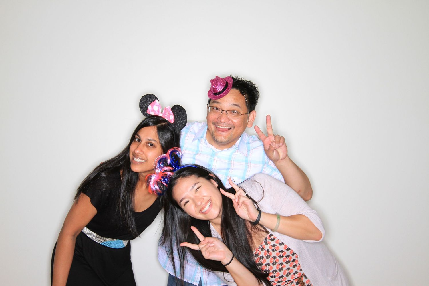 Three friends pose playfully wearing Disney Minnie Mouse ears, making peace signs and funny faces against a white background.