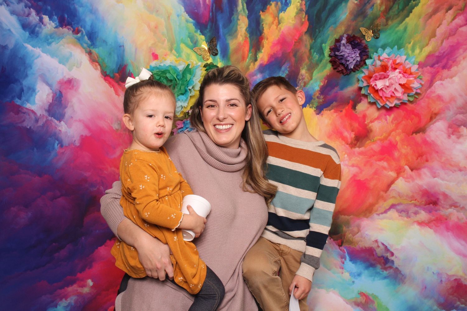 A woman smiles with two young children in front of a vibrant colorful abstract painted backdrop.