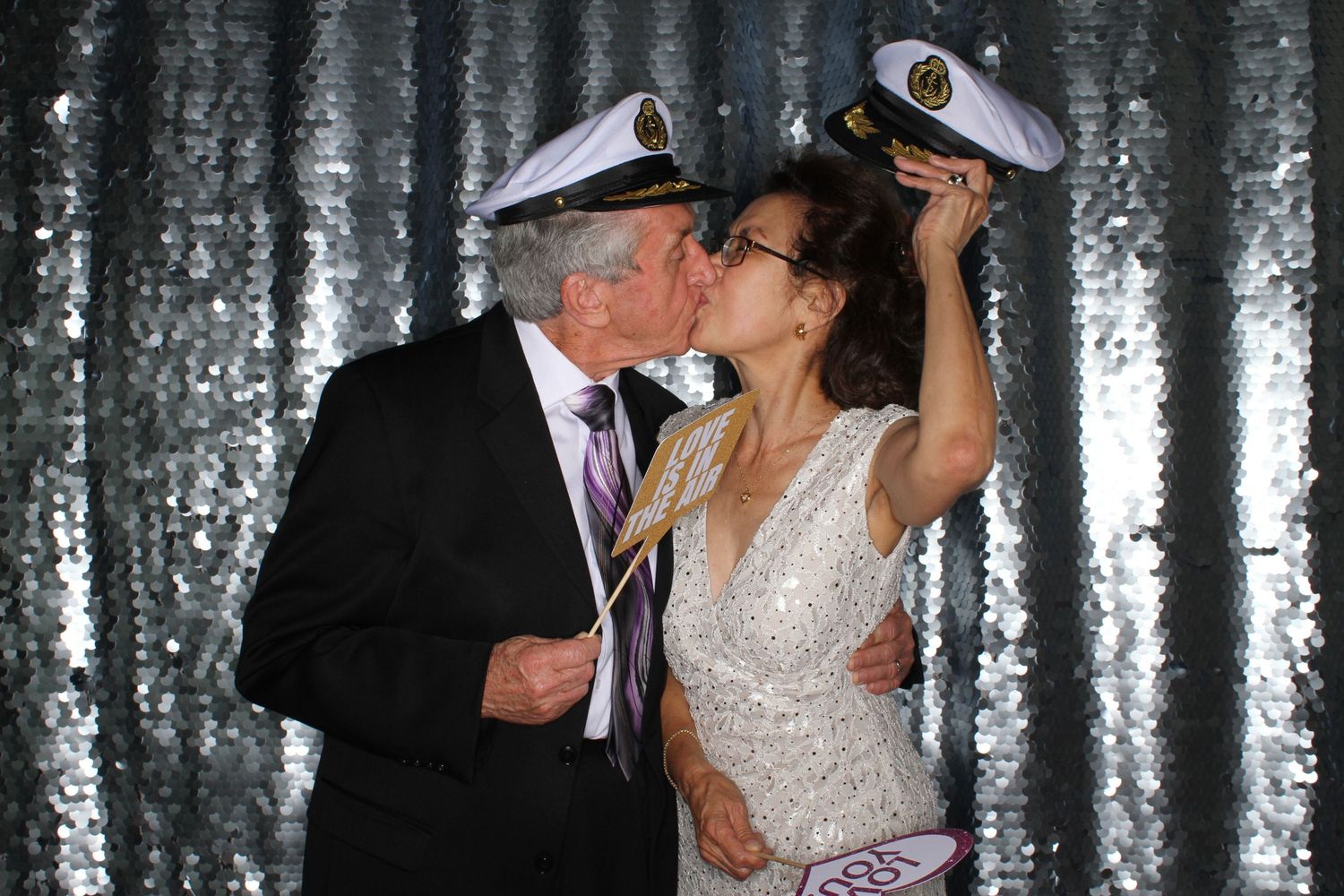 Couple kissing in photo booth wearing captain hats, woman in white dress, silver sequin backdrop.