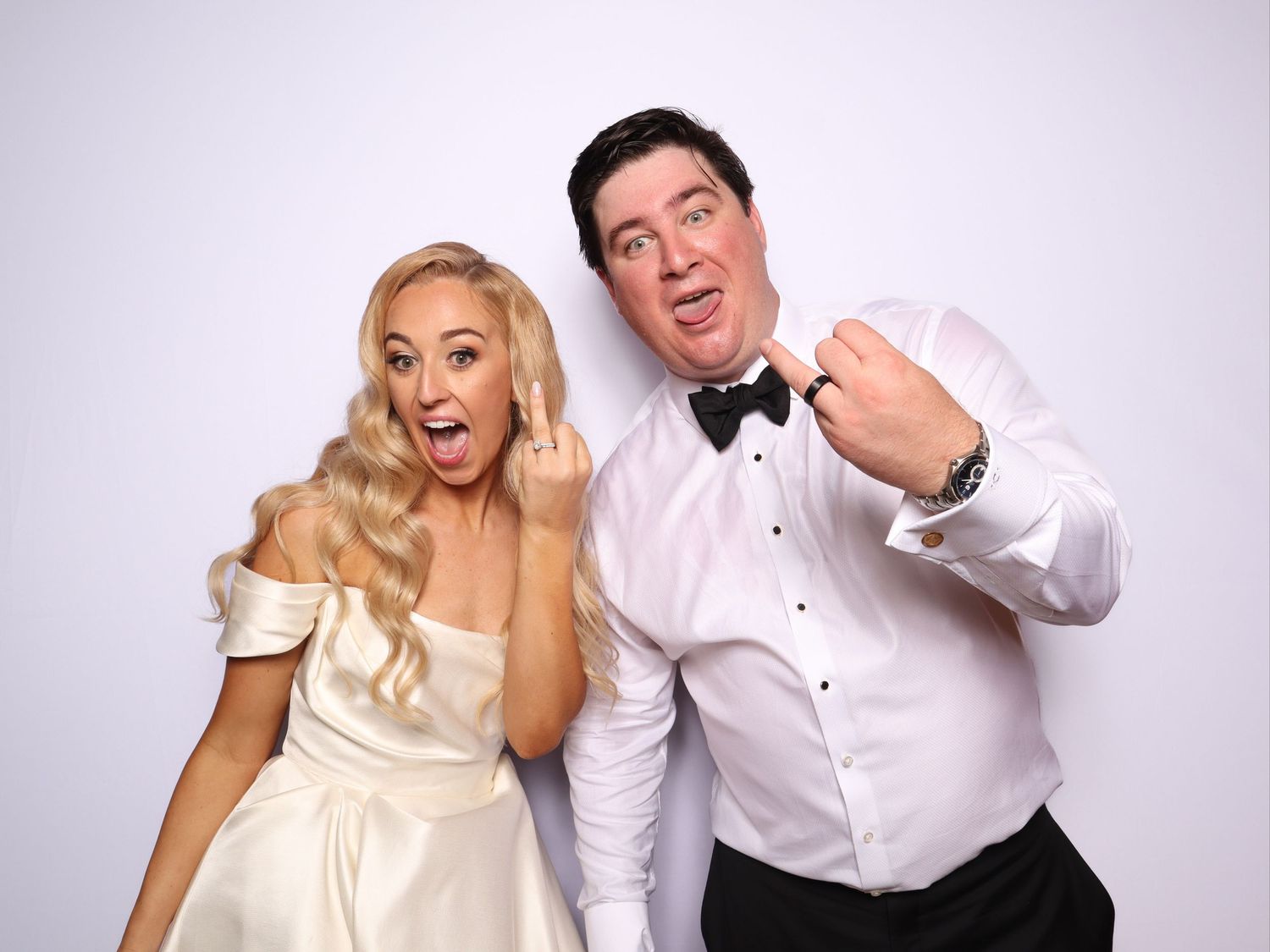 Bride and groom in formal wedding attire posing playfully showing rings at a photo booth.