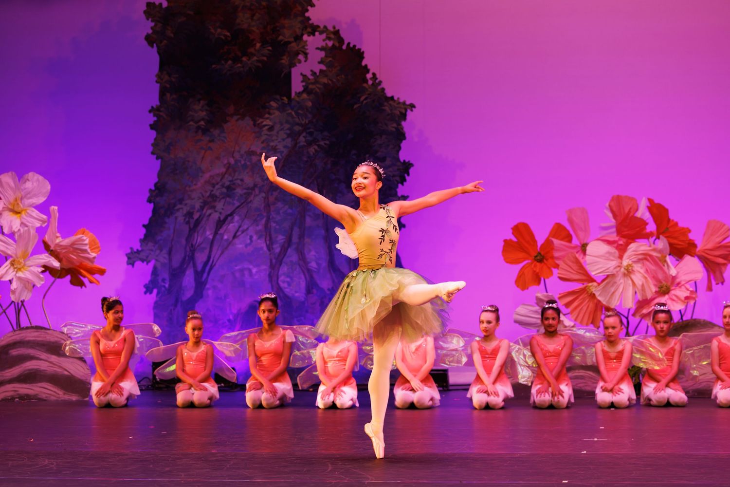 A ballerina performs on stage in a green tutu surrounded by kneeling dancers in pink costumes with floral backdrop.  Las Positas College