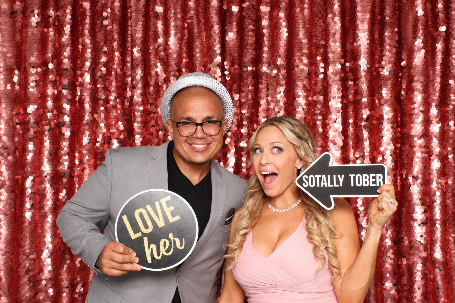 Couple posing at photo booth with sequin backdrop holding Love Her and Sotally Tober signs at a party.