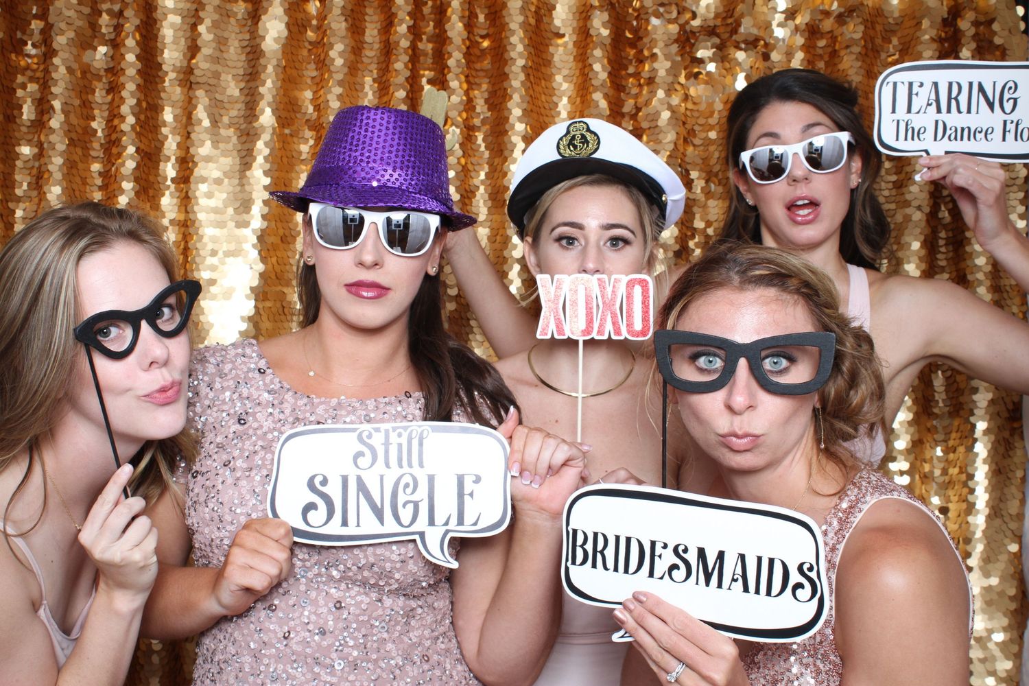 Five bridesmaids posing in a photo booth with gold sequin backdrop holding fun signs including Still Single and Bridesmaids.