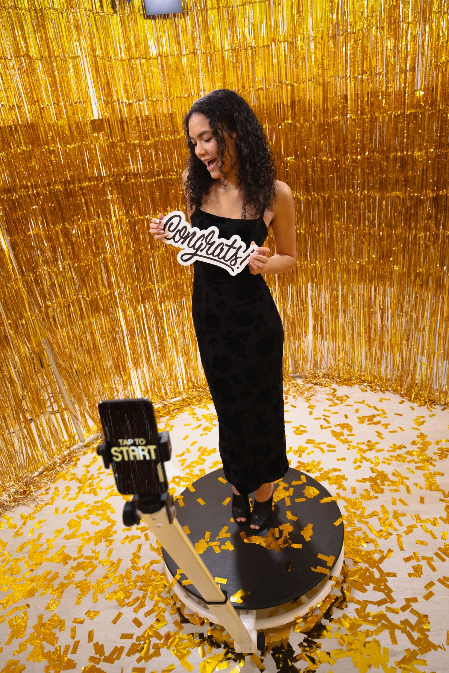 Woman in black dress holding Congrats sign on photo booth platform with gold tinsel backdrop and confetti.