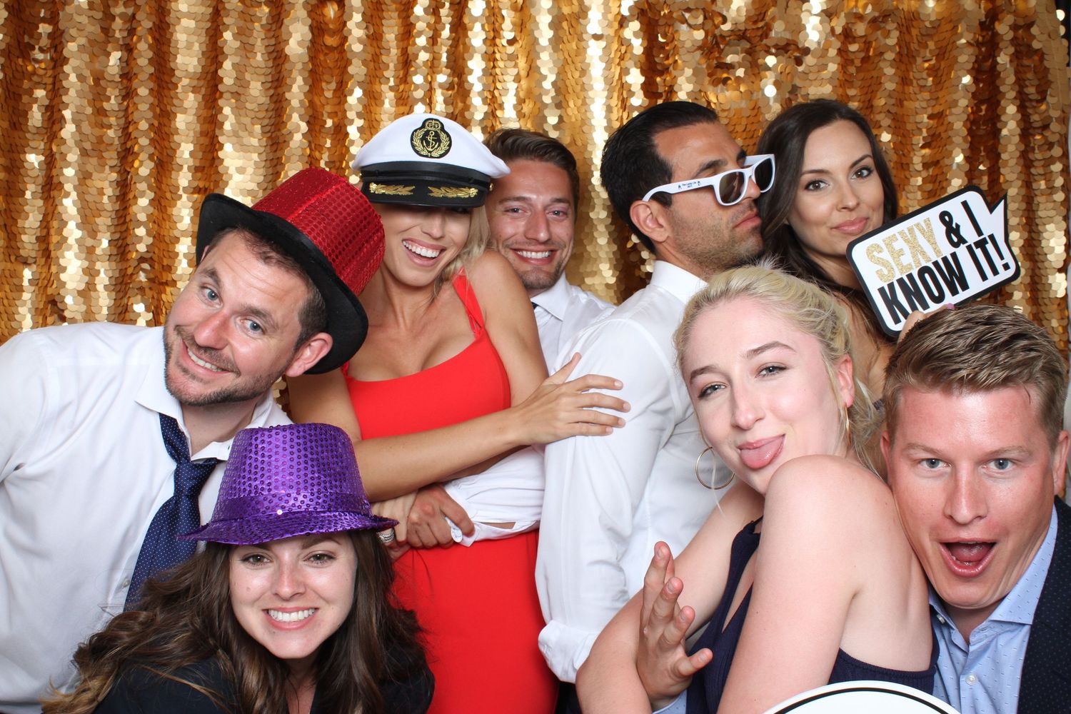 Group of friends posing in a gold sequin photo booth, wearing fun props including hats and sunglasses at a party.