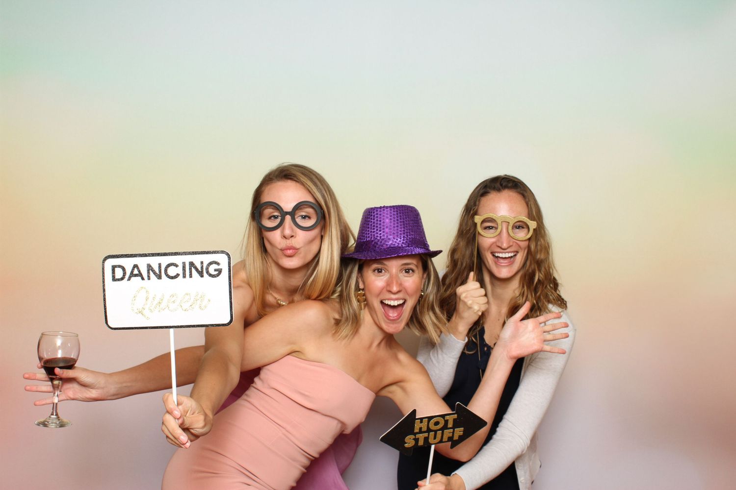 Three women pose in a photo booth with fun props including glasses, hats, and a Dancing sign, smiling and laughing together. Peacock Gap Gol