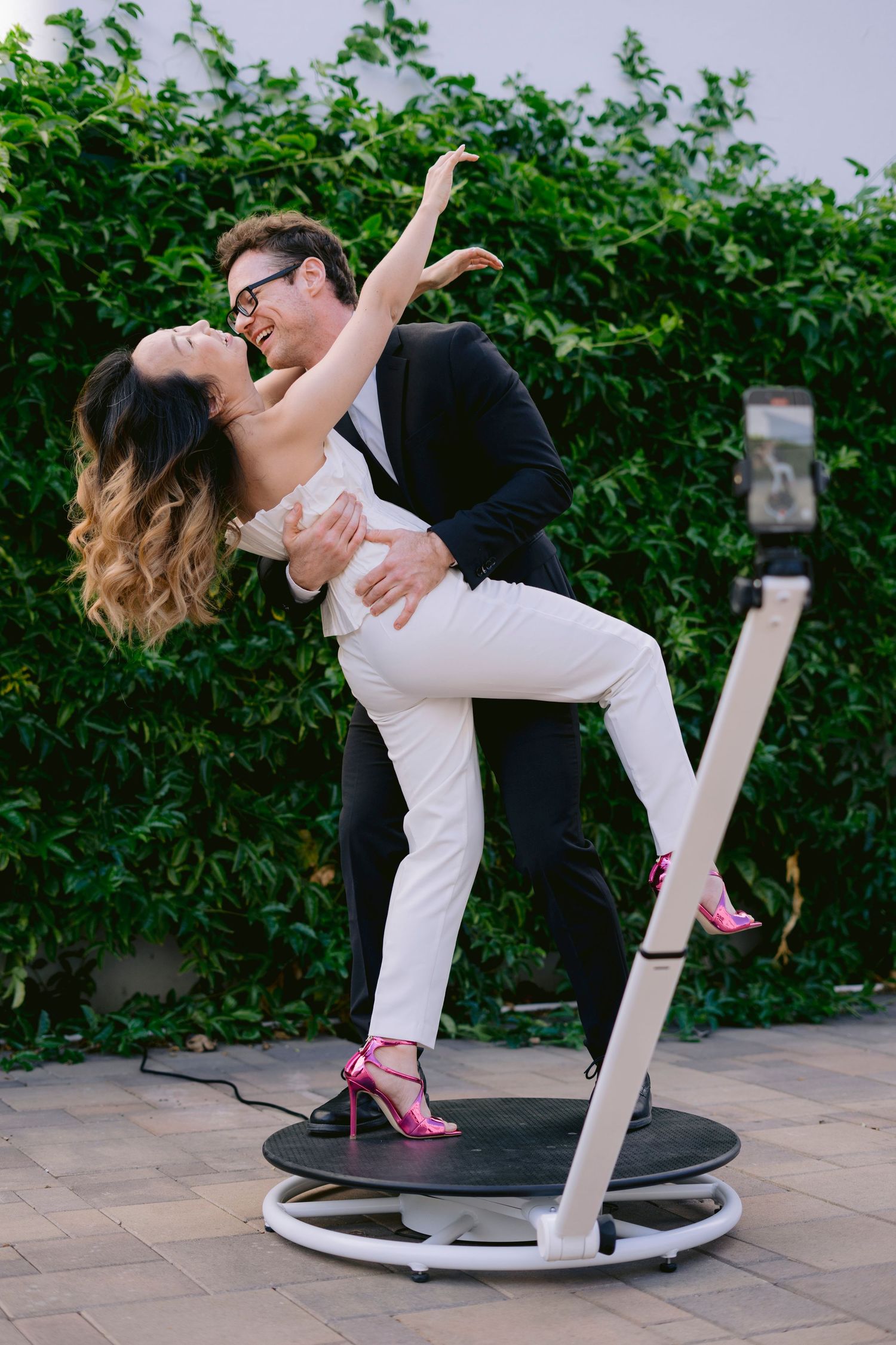 Couple in white and black outfits dancing on a rotating photo booth platform outdoors with green hedge background.