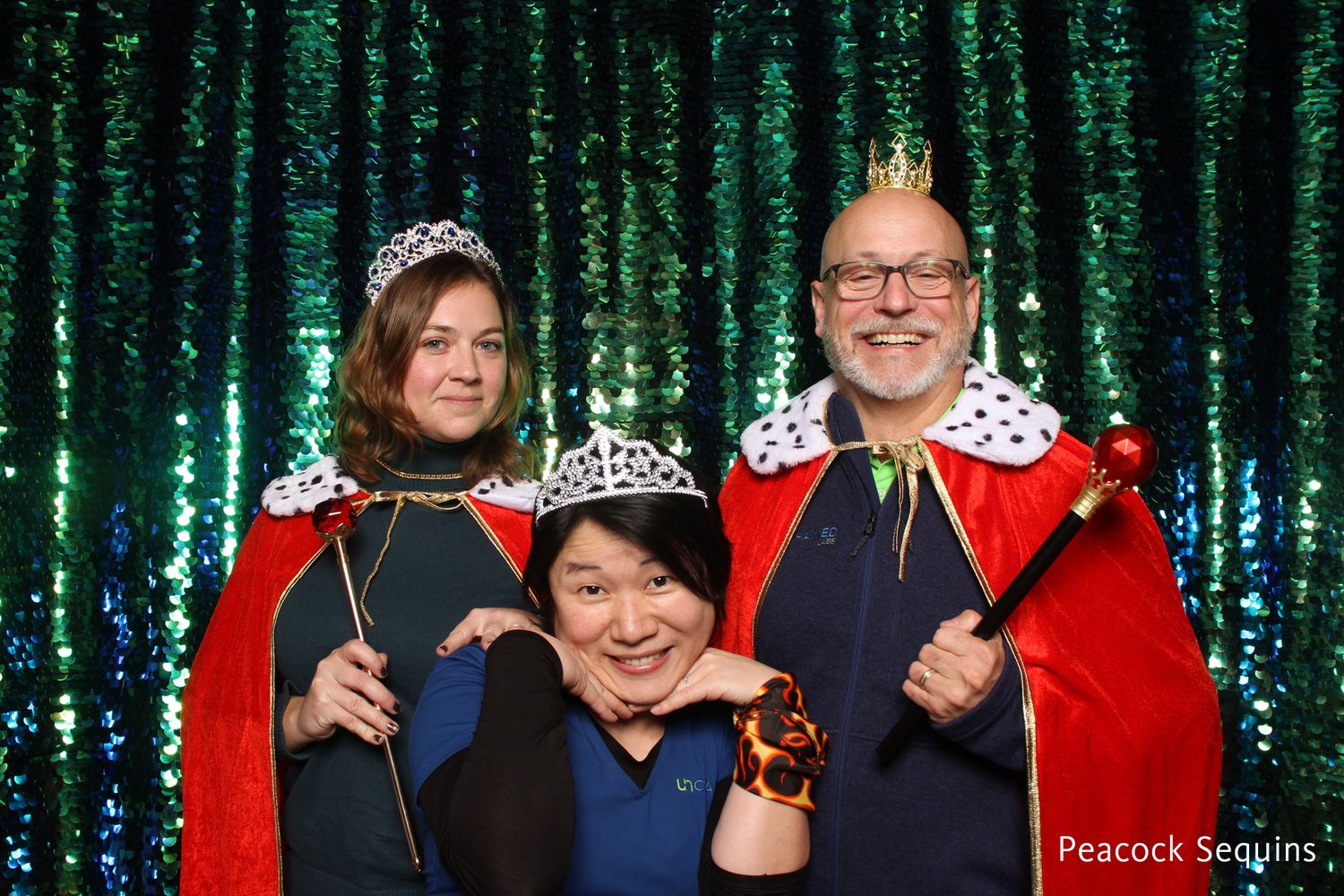 Three people in royal costumes with crowns and capes at green sequin photo booth.