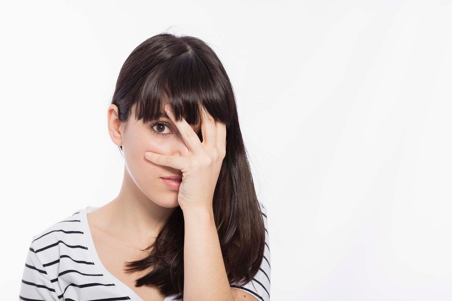 Young woman with dark hair peeking through her fingers, covering half her face, wearing a striped shirt on white background.