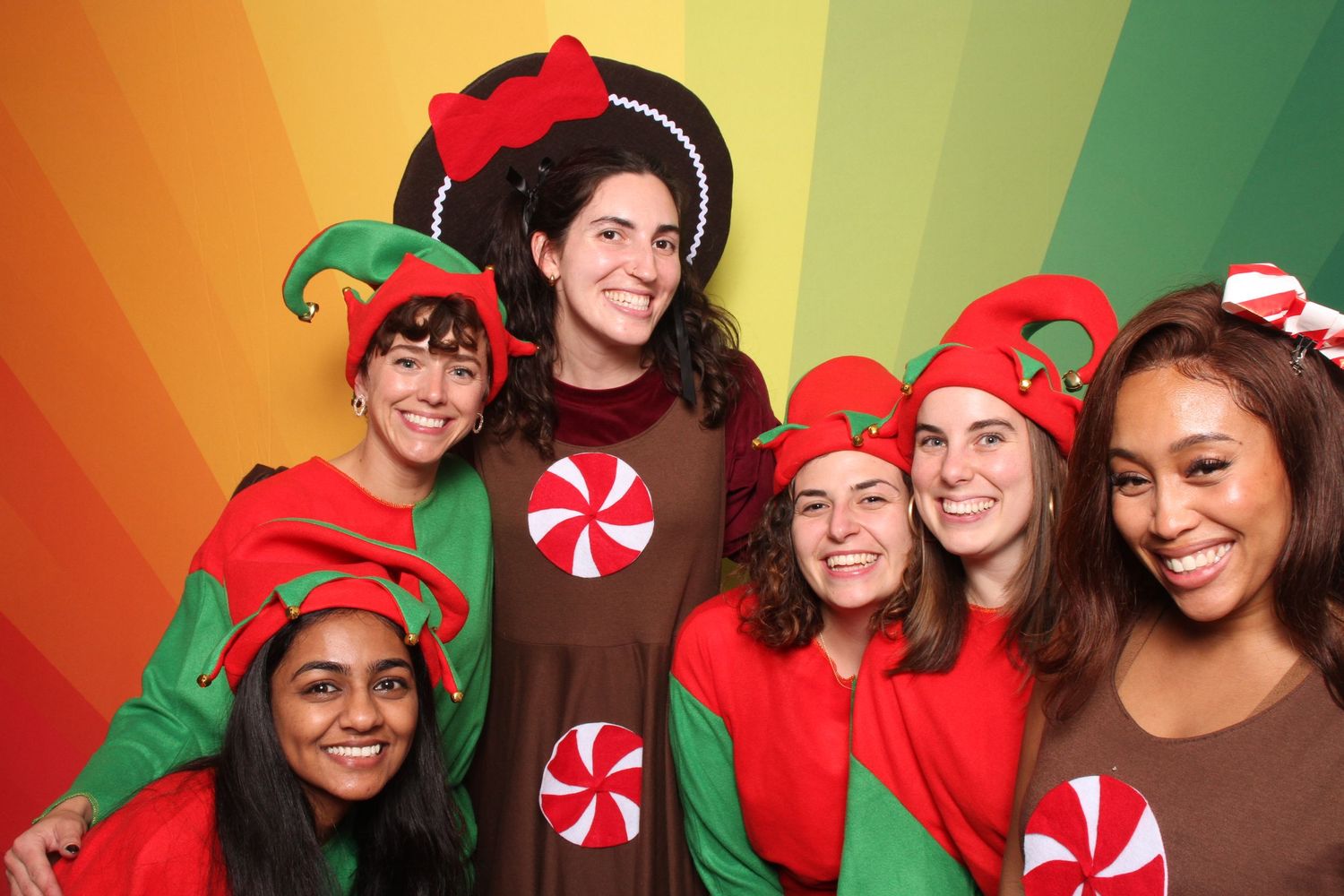 Group of six friends posing in festive Christmas elf and peppermint costumes against a colorful rainbow background.