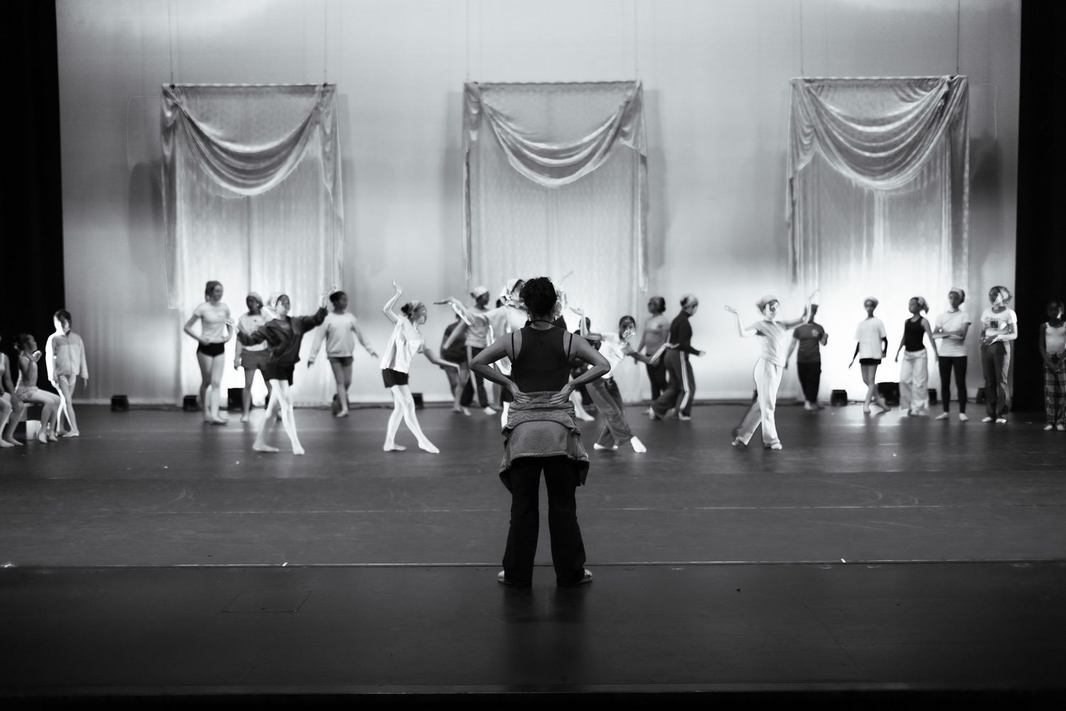 Black and white photo of a ballet instructor observing students rehearsing on a theater stage. Las Positas College