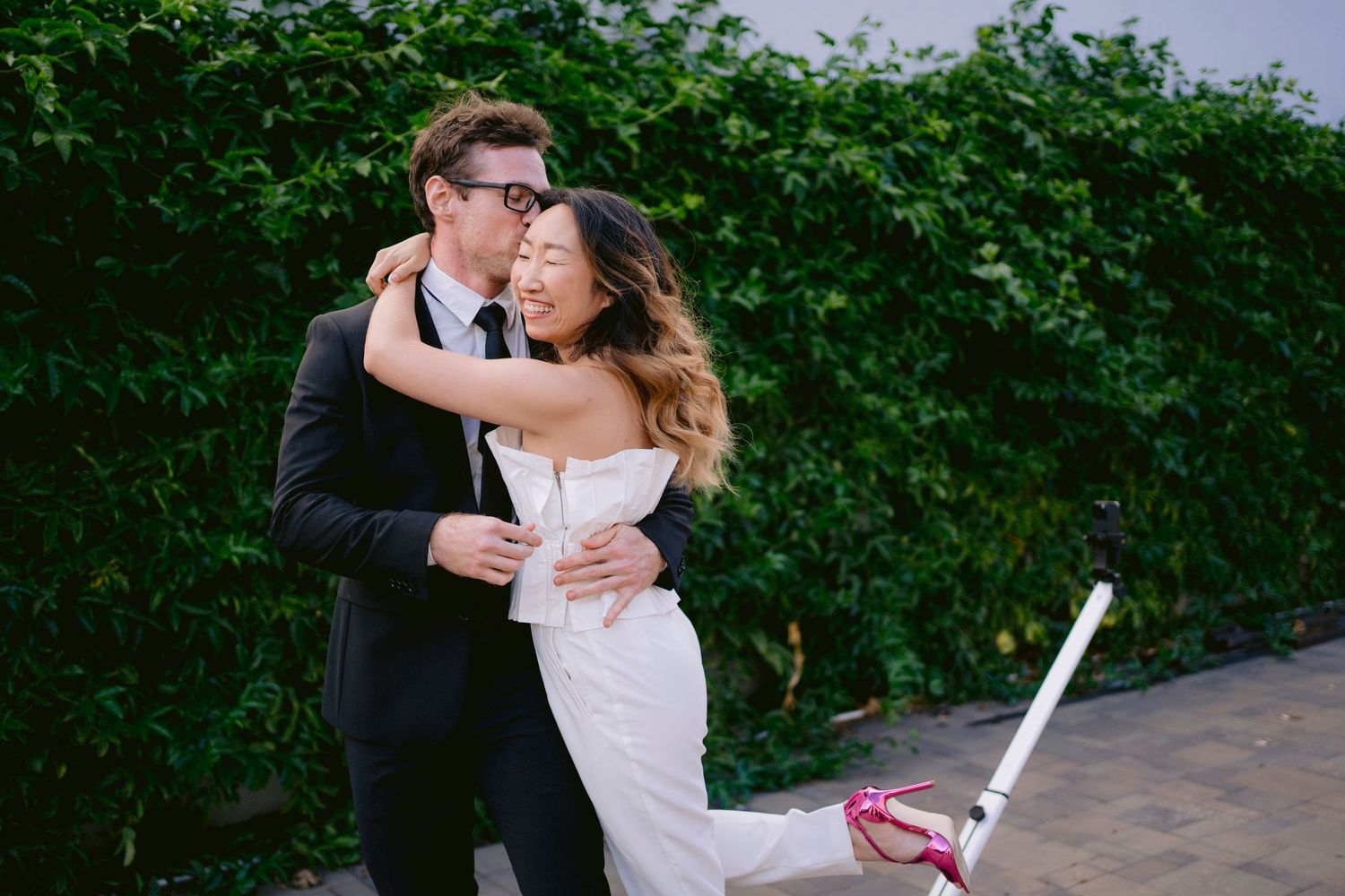 Happy couple embracing in wedding attire outdoors, bride in white gown with pink heels, groom in black suit.
