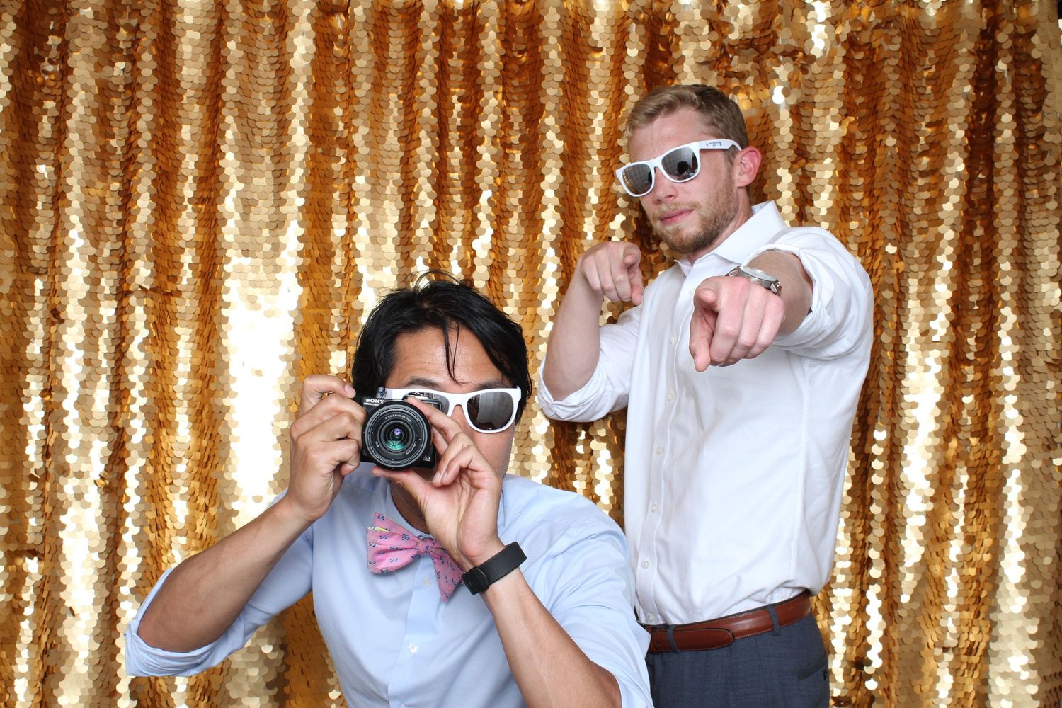Two men pose in front of gold sequin backdrop, one photographing with camera, other pointing wearing white sunglasses.