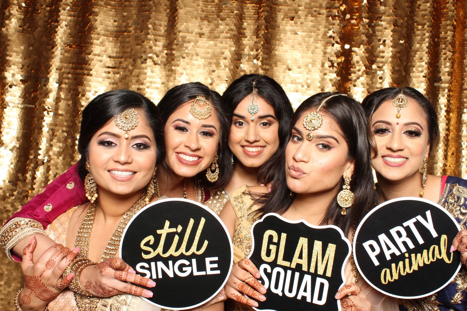 Five women in festive attire pose with fun photo booth signs reading Still Single, Glam Squad, and Party Animal.