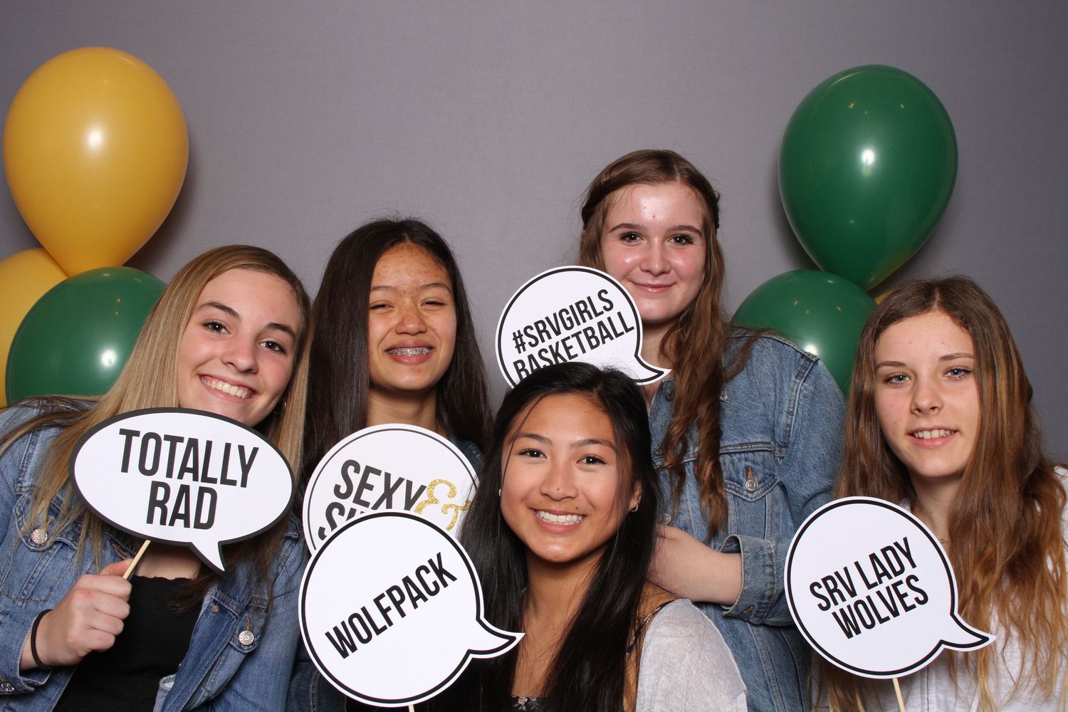 Five teen girls smile holding speech bubble props at a school basketball photo booth with green, yellow, and white balloons.