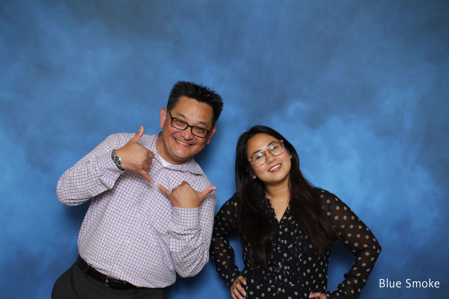 Man and woman posing with shaka signs in front of blue smoke backdrop.
