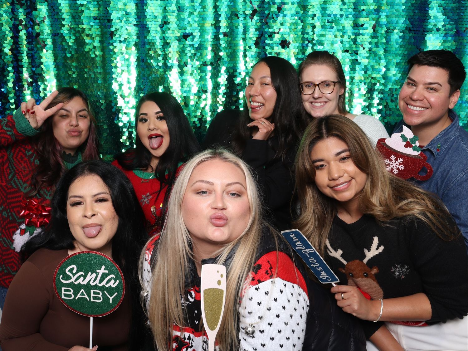 Group of friends posing at a Christmas party photo booth with a green sequin backdrop and holiday props. Gensler Holiday Party