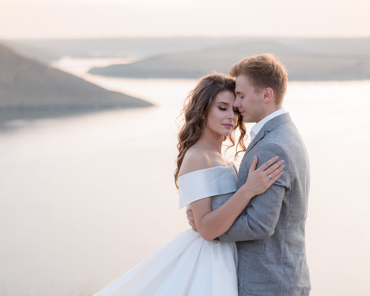 A bride and groom embrace romantically outdoors with a scenic lake and hills in the soft background light. Audio guestbook audiogram.