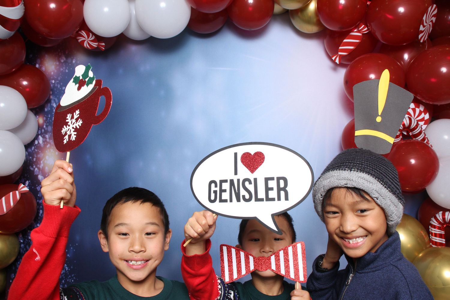 Three children pose at a festive Christmas photo booth with balloon arch, holding an I Love Gensler sign and holiday props.