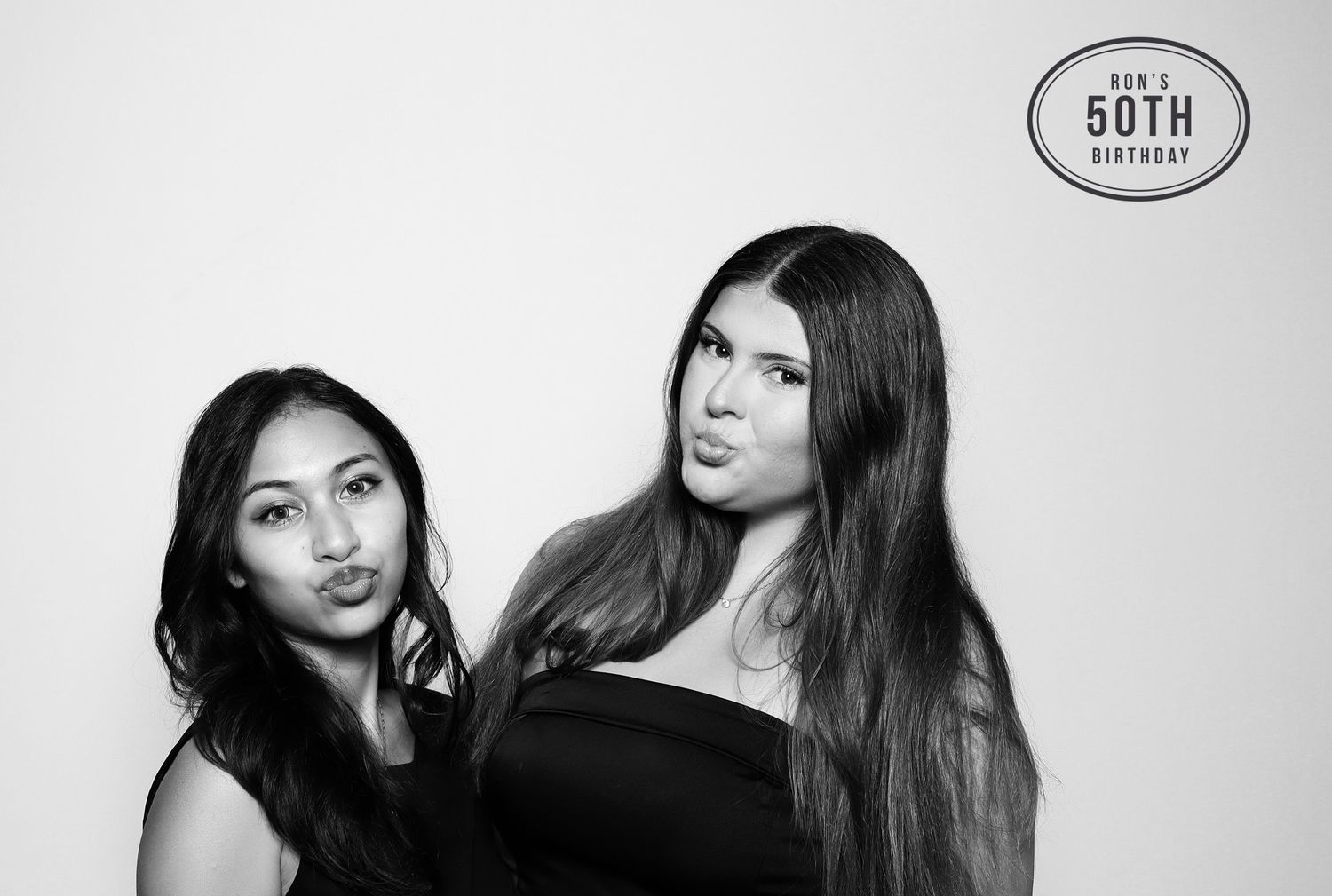 Two women pose playfully in black outfits at a 50th birthday photo booth, captured in black and white. Industrious Walnut Creek.