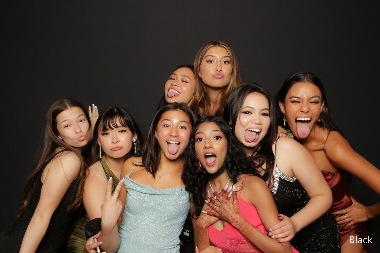 Seven young women in formal dresses posing playfully in a photo booth with black background.