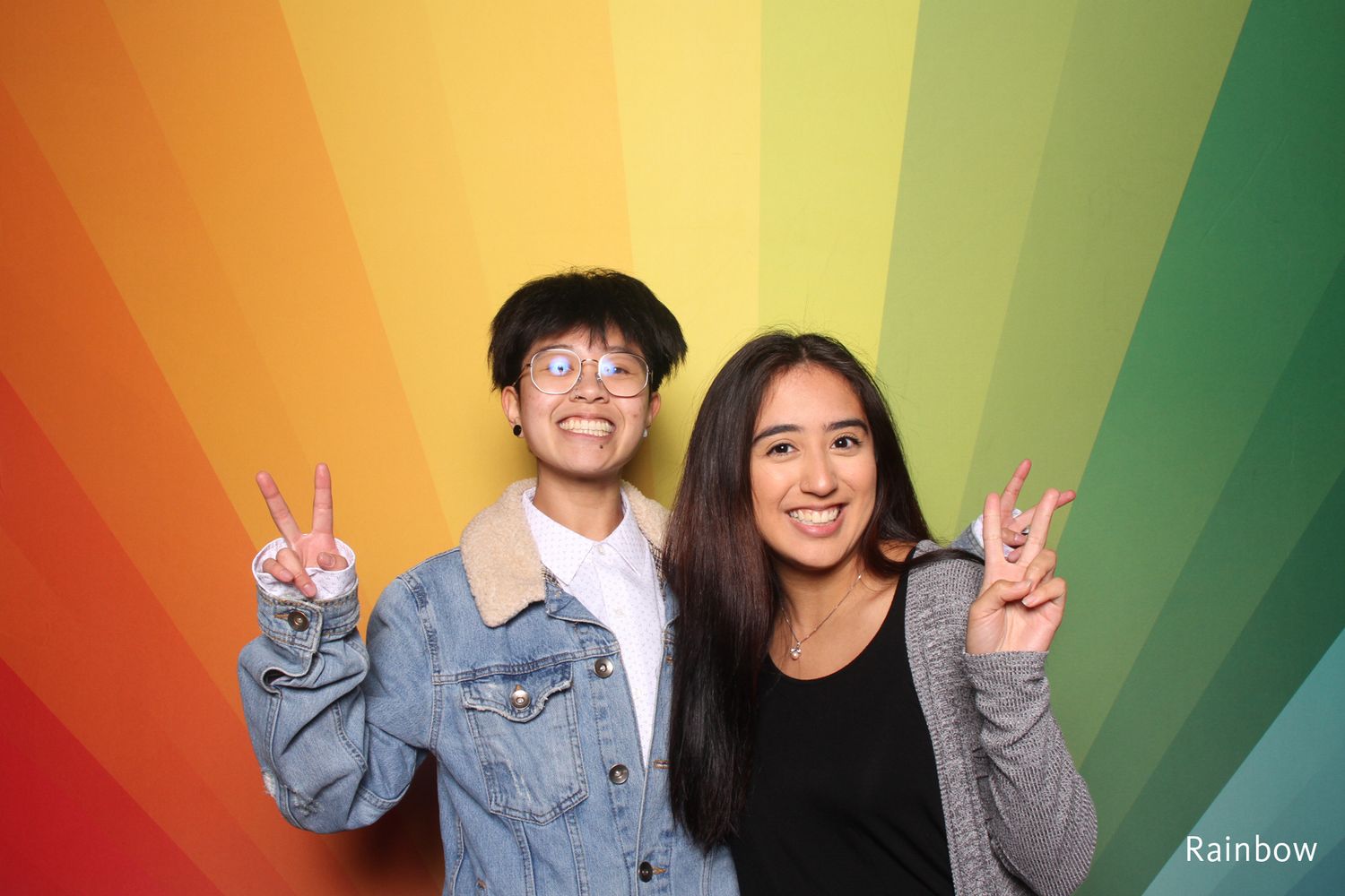 Two young people flashing peace signs in front of colorful rainbow sunburst photo booth backdrop.