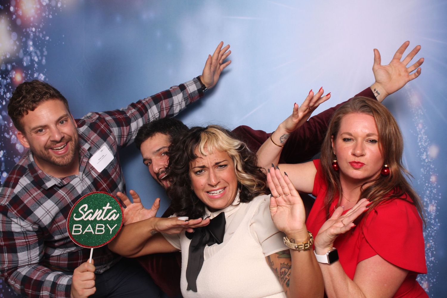 Four friends posing playfully at a Christmas photo booth, holding a Santa Baby sign with festive decorations.