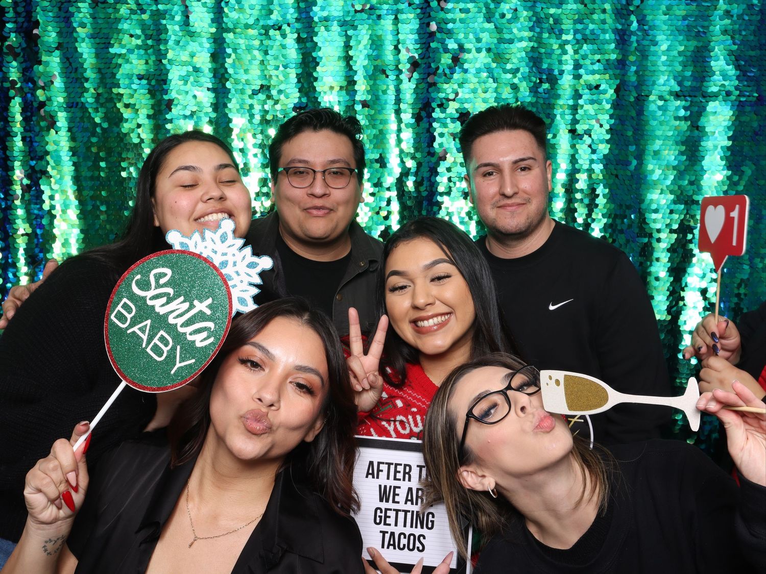 Group of six friends posing in a festive photo booth with green sequin backdrop, holiday props, and fun expressions. Gensler Holiday Party
