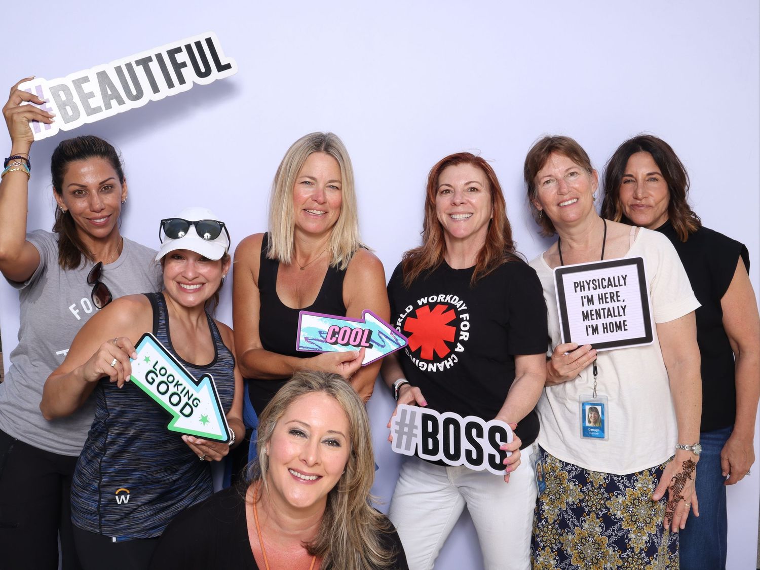 Group of seven women posing with fun signs including Beautiful, Boss, and Cool at a photo booth event.